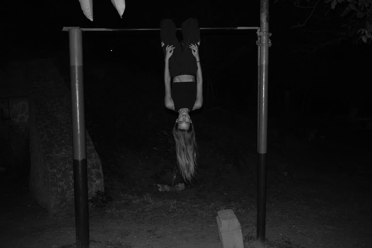 Anastasiia Koval inverted on an outdoor training bar at night, in black and white.